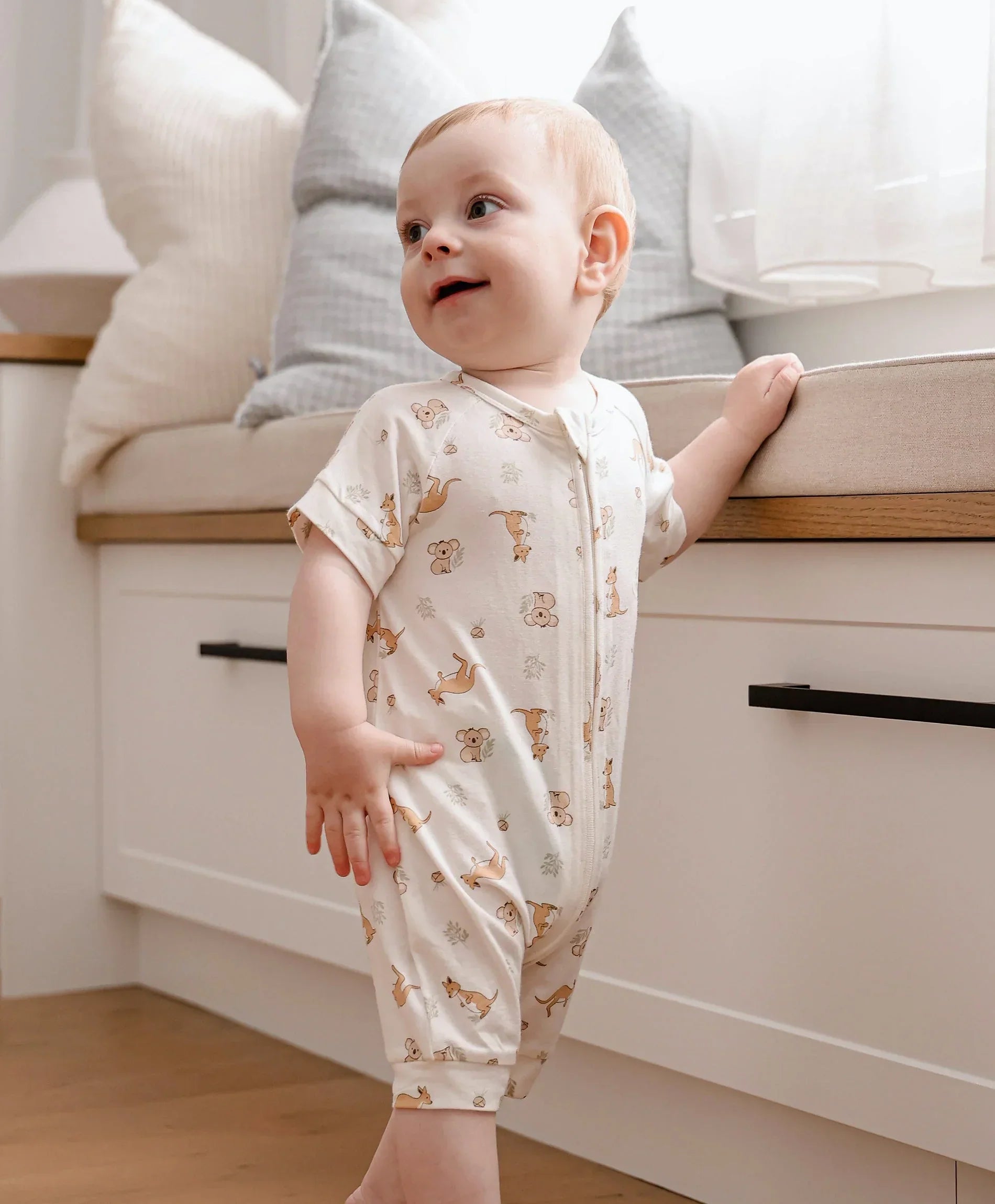 Baby in a printed short-sleeve organic cotton romper, standing and holding onto a bench in a bright home setting
