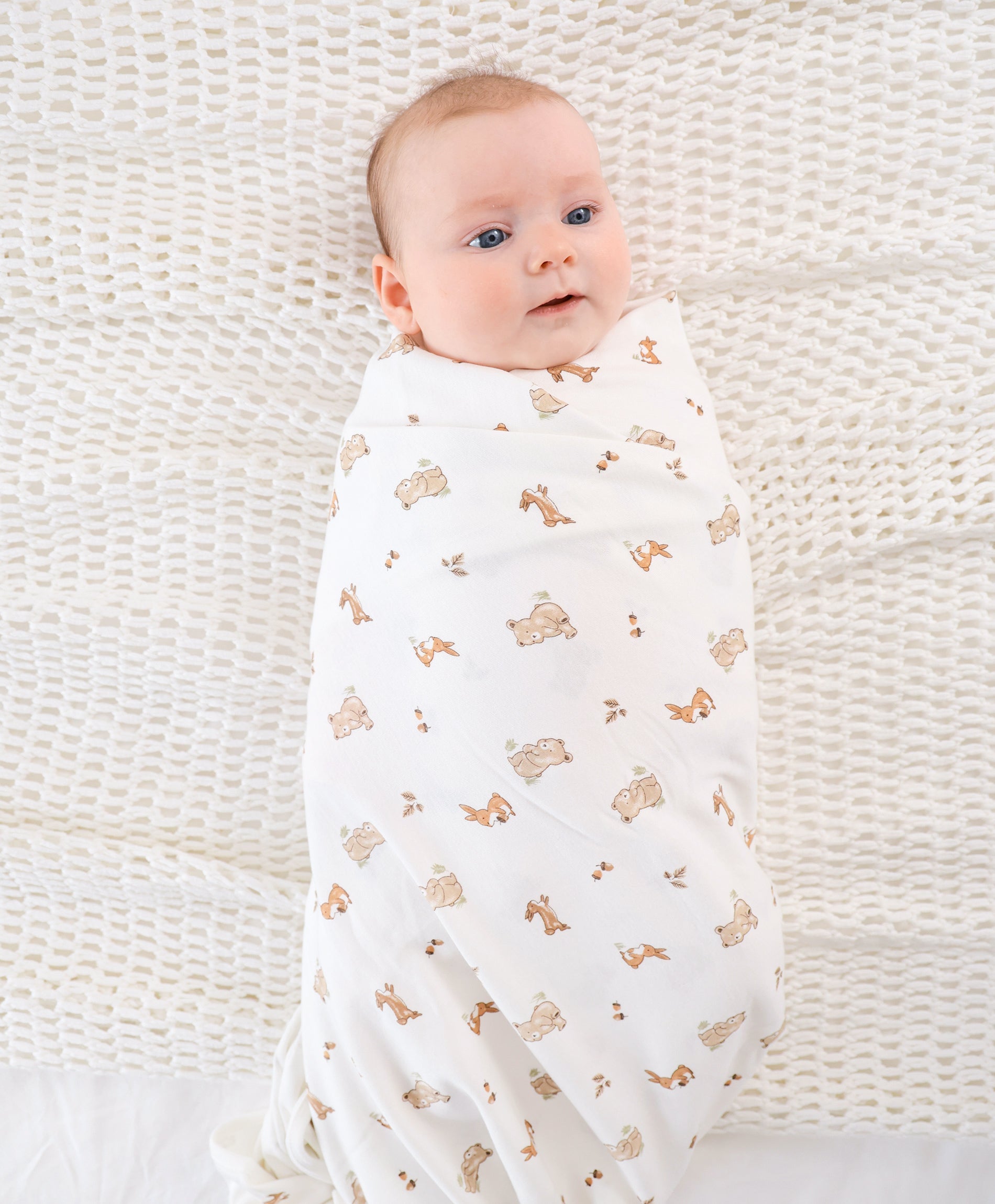 Newborn baby swaddled in a white muslin wrap with small bear and bone prints, lying in a white crib